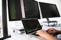 A developer writes code on a laptop in front of multiple monitors in an office setting.