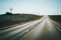 A peaceful rural highway stretching into the distance, flanked by telephone poles and open fields under a clear summer sky.