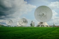 Array of large satellite dishes in a lush green field under a cloudy sky.