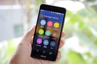 A hand holding a smartphone displaying home management apps on the screen.