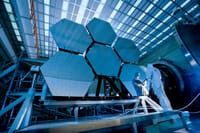 Engineers in protective suits work on telescopic mirrors in a high-tech lab.