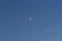 A crescent moon and airplane contrail against a clear blue sky, creating a serene contrast.