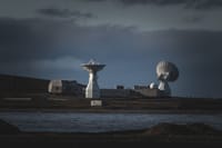 A remote satellite station with large dishes stands under a moody dark sky by the water.