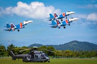 Three aerobatic jets performing a formation flight above a helicopter on a clear sunny day.
