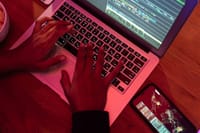 Hands typing on a laptop with code on screen, smartphone nearby at night, indoor setting.