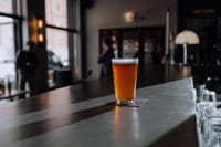 A glass of craft beer with foam sits on a bar counter in a dimly lit modern bar.