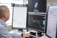 Stock trader analyzing financial graphs on multiple computer monitors in an office setting.
