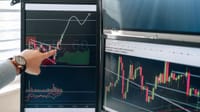 Person pointing at stock market graphs on dual monitors in a modern workspace.