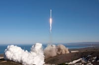 A rocket launches over the ocean, leaving behind smoke and fire trails. Scenic backdrop with clear skies.