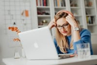 Blond woman wearing eyeglasses and headphones, working on a laptop indoors at a desk.