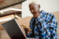 Young serious African American female in blue checkered shirt looking at screen of netbook while texting message on mobile phone
