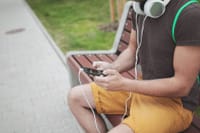 A young adult seated on a park bench, listening to music on headphones connected to a smartphone.