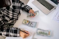 A woman in a plaid coat counts American dollars at a desk with a notebook and laptop.