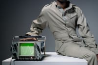 A person in coveralls sits beside an oscilloscope displaying a waveform in a studio setting.