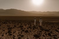 Astronauts in space suits explore a rocky, desert-like terrain resembling Mars at sunset.