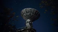 From below of blue starry sky over radio telescope and trees with leaves