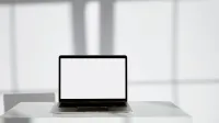 Blank laptop screen on a modern white desk in a bright, sunlit room.