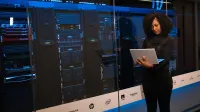 Una ingeniera que usa una computadora portátil mientras monitorea los servidores de datos en una moderna sala de servidores.