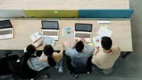 Aerial view of four colleagues working with laptops and documents at a shared desk.