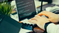 Close-up view of a developer typing code on a keyboard with a computer screen showing scripts.