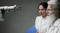 Two scientists wearing lab coats and goggles analyzing a robotic arm in a laboratory setting.