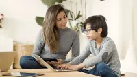 A mother and her son using a laptop and tablet together in a cozy home setting.