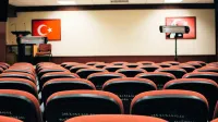 Una sala de conferencias vacía con asientos rojos, un escenario y banderas turcas, lista para una reunión o presentación.