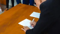 A close-up of a businessman signing official documents at a wooden desk.