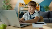 Young boy using a laptop for online learning at home with study materials nearby.