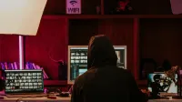 Person in a hoodie editing videos on multiple screens in a dimly lit space with neon lighting.