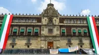 Front view of the Mexican National Palace in Mexico City with large Mexican flags displayed, a bright sky overhead.