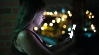 A young woman engaging with her smartphone on a balcony at night, surrounded by city lights.