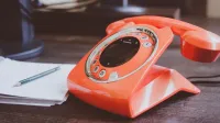 Un teléfono giratorio naranja de estilo retro se encuentra en un escritorio de madera junto con un lápiz y un bloc de notas.