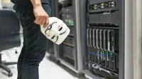 Persona que sostiene una máscara anónima cerca de servidores, insinuando la seguridad cibernética y hackeando temas.