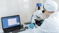 Scientist in lab coat using microscope and laptop in a laboratory setting.