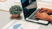 Hands typing on a laptop at a desk with an insurance paper and plant, suggesting a work environment.