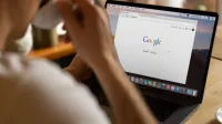 Close-up of an adult drinking coffee and browsing Google on a laptop indoors.