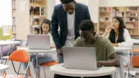 Students learning in a classroom setting with a teacher assisting and laptops on desks, creating an interactive education environment.