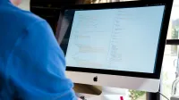 A programmer in a blue shirt coding on an iMac. Perfect for technology or work-related themes.