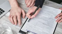 Close-up image of two people signing an insurance policy document on a wooden desk.