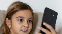 A young girl with long brown hair smiles while using a smartphone indoors.