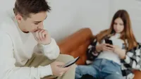 Two teenagers sitting casually on a couch, absorbed in their smartphones indoors.