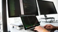 A developer writes code on a laptop in front of multiple monitors in an office setting.