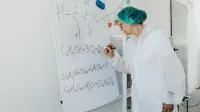 Anciana en bata de laboratorio escribiendo ecuaciones químicas en una pizarra en el interior.