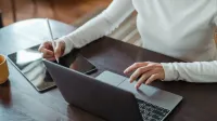 From above of crop anonymous businesswoman in white sweater working at table using tablet with stylus and typing on laptop