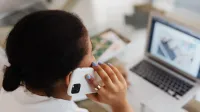 Mujer sosteniendo un smartphone a la oreja mientras trabaja en una computadora portátil. Comunicación moderna y multitarea.