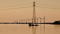 Silhouette of wind turbines and power lines over water during sunset, reflecting sustainable energy.