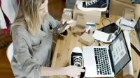 Mujer fotografiando zapatos para la venta en línea en el espacio de trabajo del hogar con laptop y paquetes.