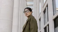 Portrait of a stylish woman in eyeglasses, olive coat, against modern architectural backdrop.