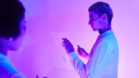 Two individuals engaging with futuristic transparent touch screens in a vibrant purple-lit studio.